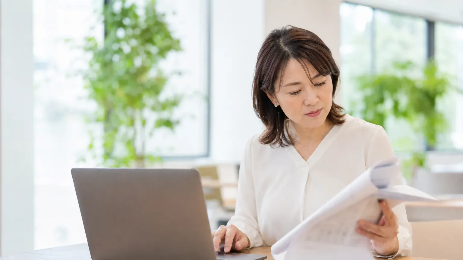 紙の帳票の束をめくりながら、正面のノートPCで内容を確認している40代女性の担当者