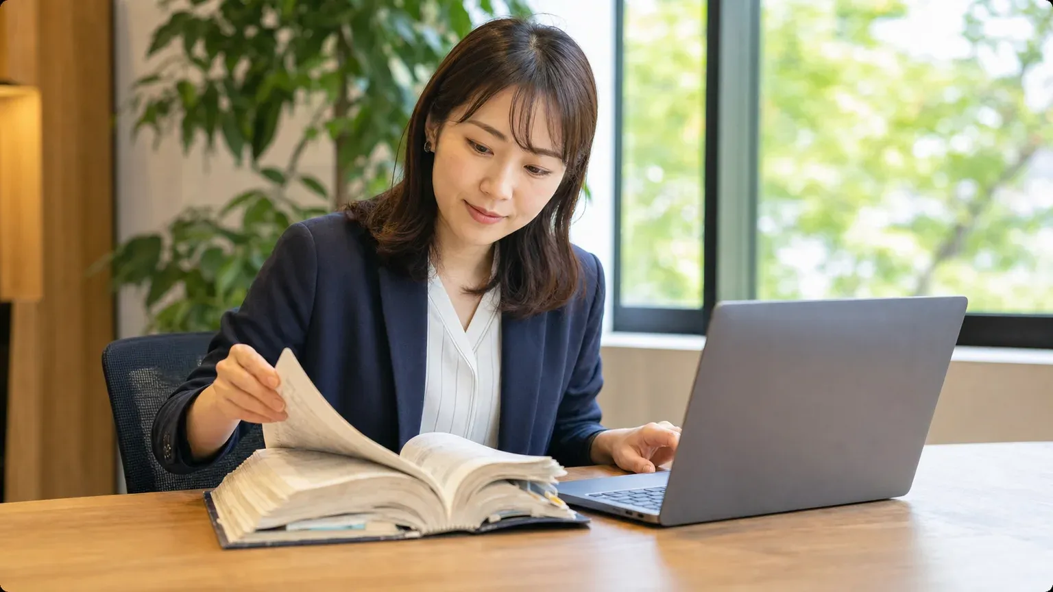 窓際のデスクで分厚い紙の業務マニュアルをめくりながらノートPCで現行の基幹システムを確認する30代の情報システム担当者