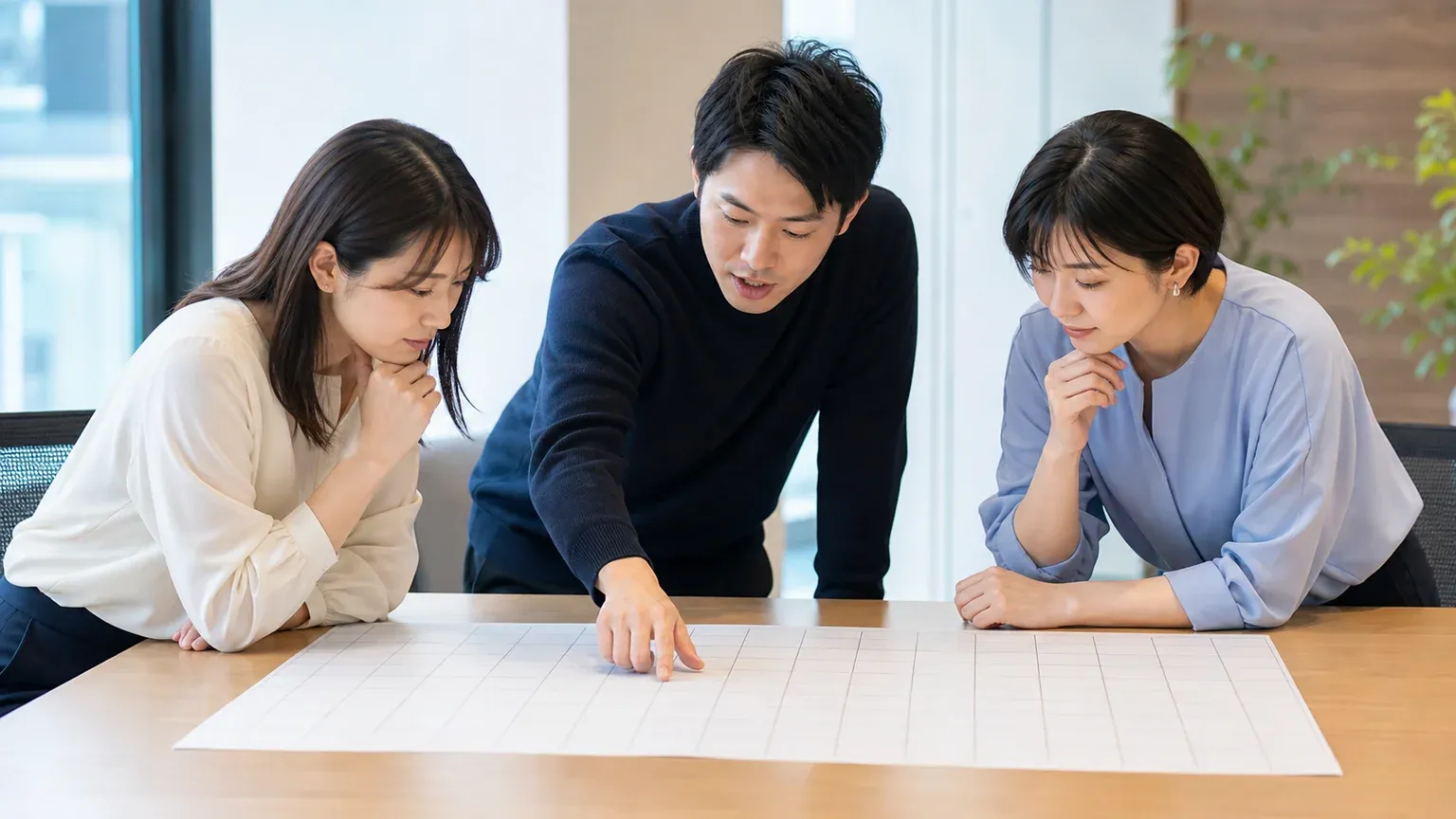 会議室で20代から40代の男女3名が年間の予定表を前に、いつ判断するかの期日を相談している引きの場面