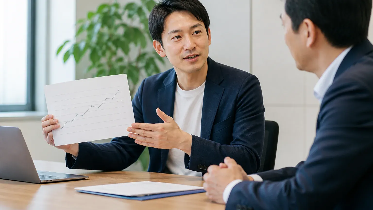 明るい会議室で30代男性の担当者が推移をまとめた資料を手元に示しながら、斜め向かいの相手に現状を説明している場面