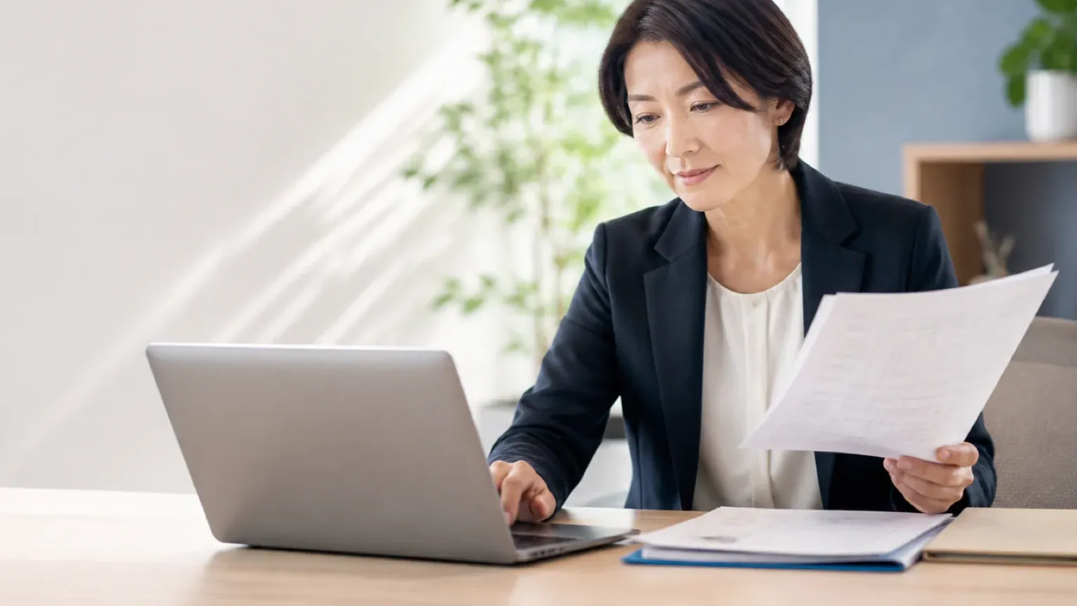明るいオフィスで40代女性のエンジニアが、現行システムの紙資料とAIが生成した設計情報を突き合わせ、内容が合っているかを確認している様子