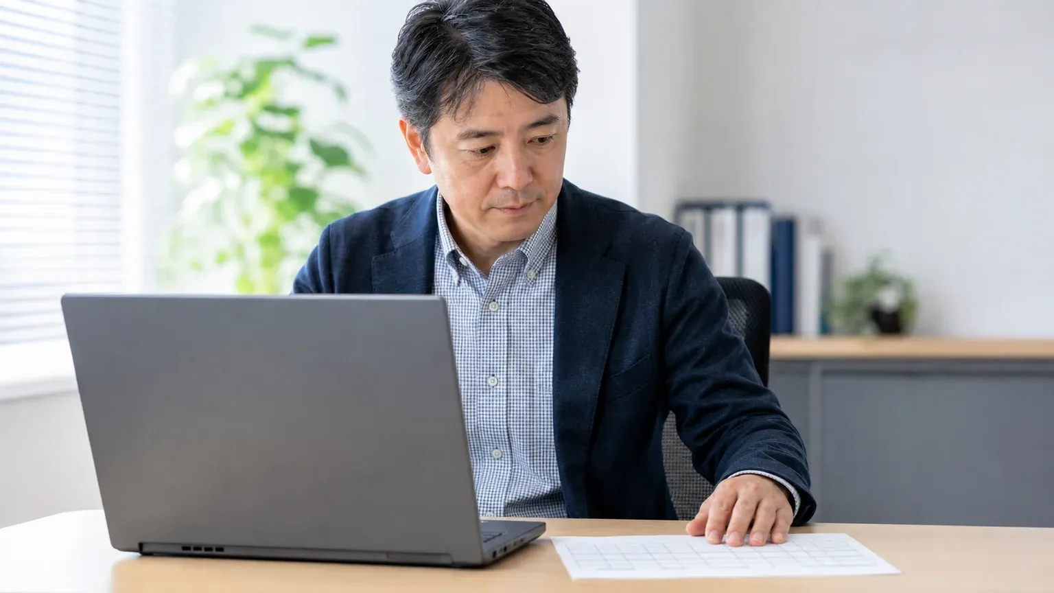 明るいオフィスで40代男性の現場担当者が画面を操作しながら、手元の紙資料と見比べて実際の業務が回るかを確認している寄りの場面