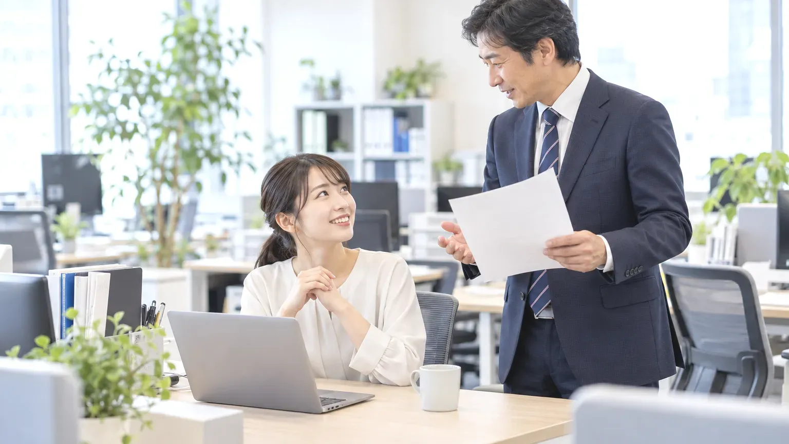 自席にいる担当者のところへ同僚が立ち寄り、手に持った紙を見せながら契約の条件を相談している場面