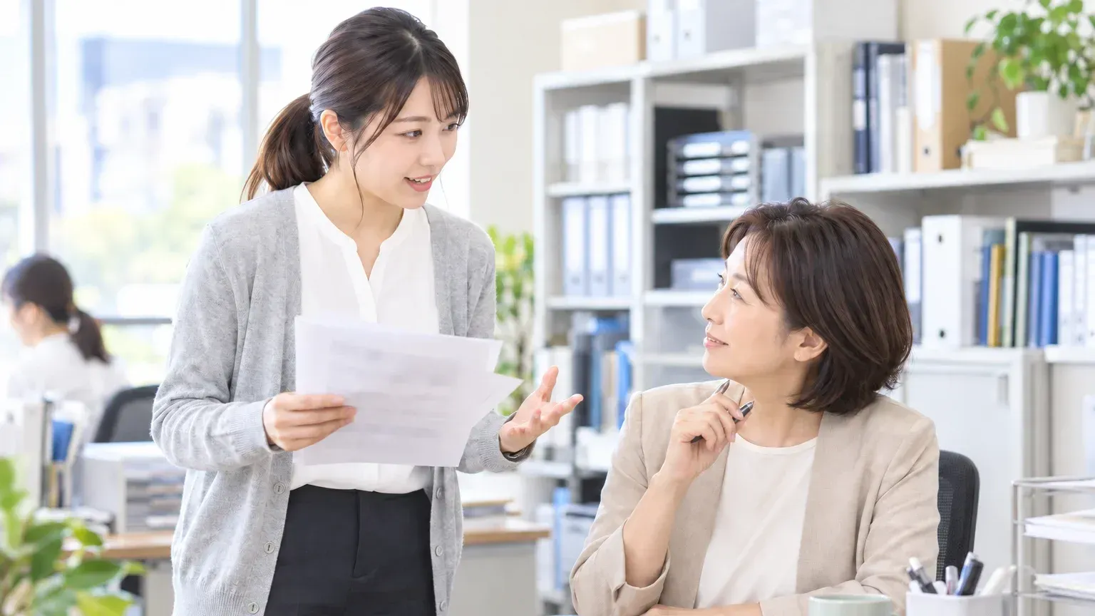 印刷した開発会社の一覧を手にした担当者が、上司の席を訪ねて依頼先の候補を相談している様子