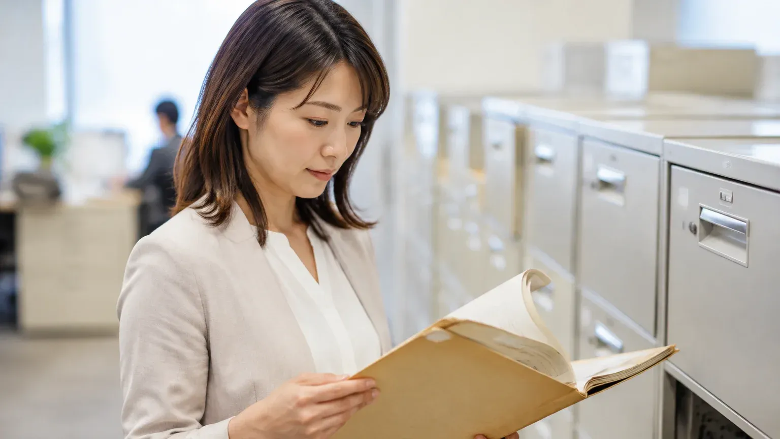 書類キャビネットの前で、担当者が古い紙のファイルを1冊抜き出して中身を確かめている場面