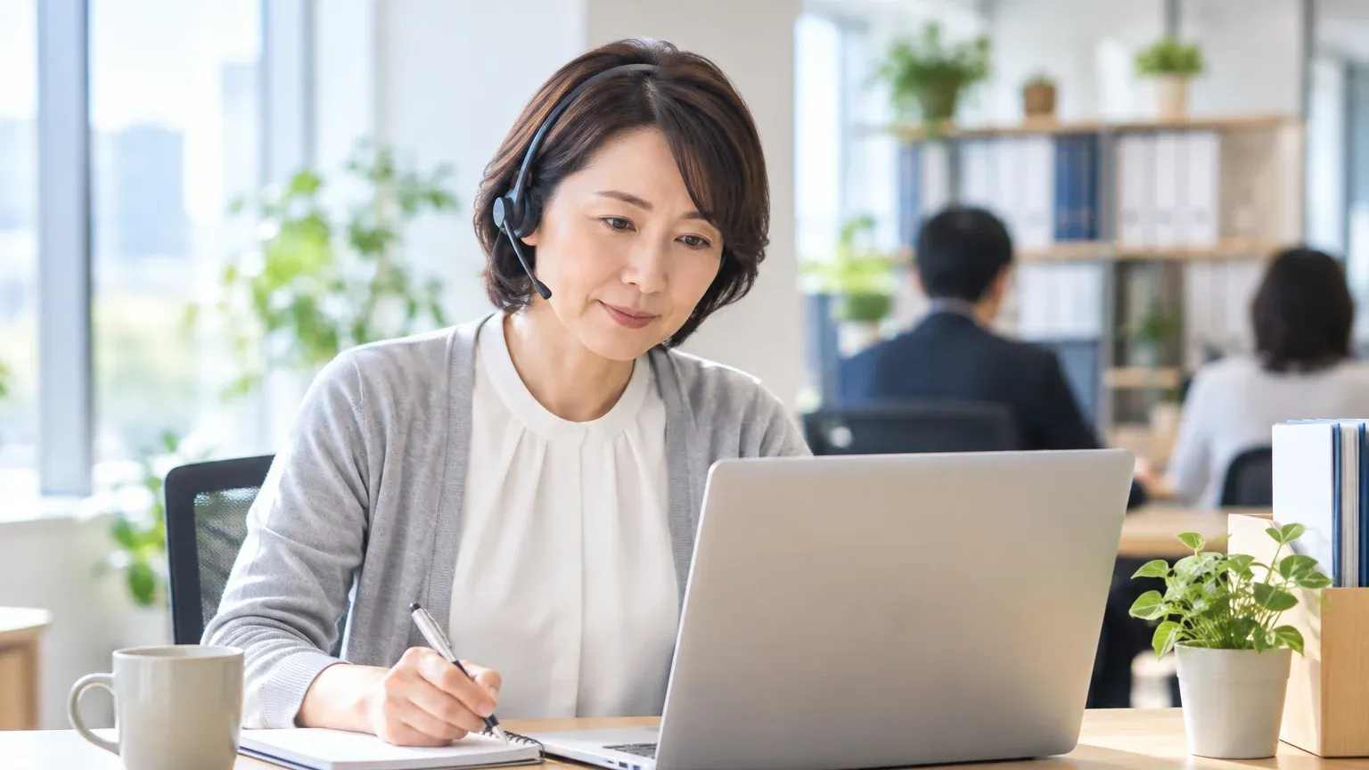自席でヘッドセットを着けた担当者が、オンライン会議で委託先の説明を聞きながらノートにメモを取っている様子