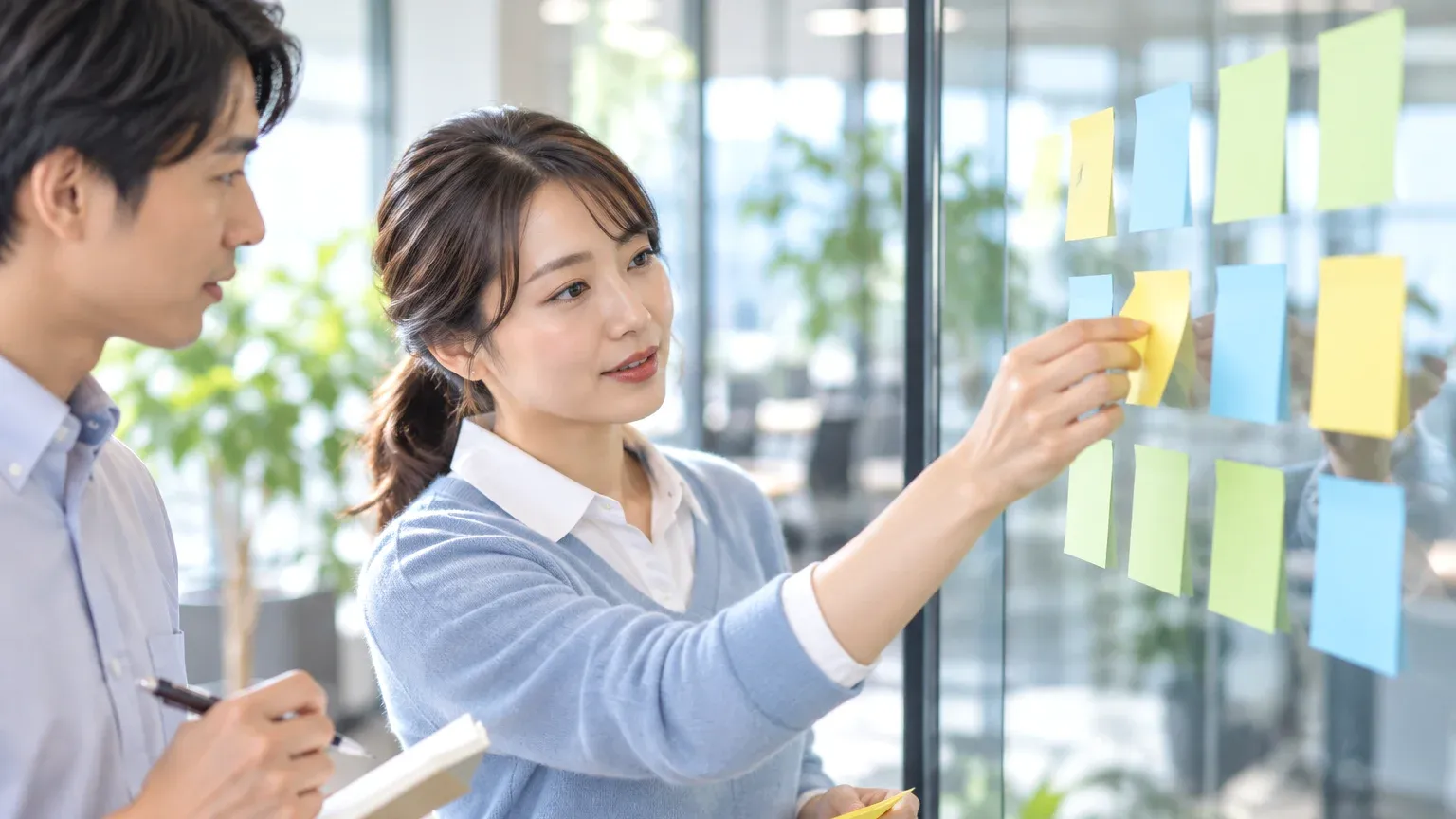 廊下のガラス壁に工程ごとに貼ったふせんを、担当者が1つ前の列へ貼り直している様子
