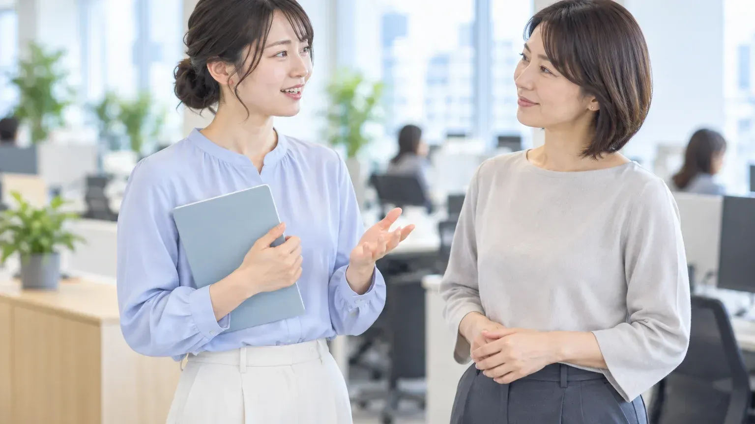自席のそばで2人の担当者が立ったまま、何から手をつけるかを短く話し合っている場面